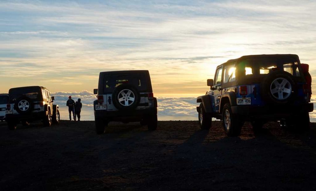 Three rental cars from Big Island Hawaii stand parked in front of a stunning sunset backdrop, with vibrant hues painting the sky and fluffy clouds adding texture to the scene