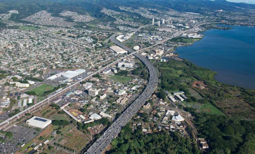 A bird's-eye view captures the sprawling cityscape of Oahu, with high-rise buildings, long streets that's coming into the view of the camera, green nature and ocean shore along the landscape. It's showcasing the urban life of Hawaii's capital.