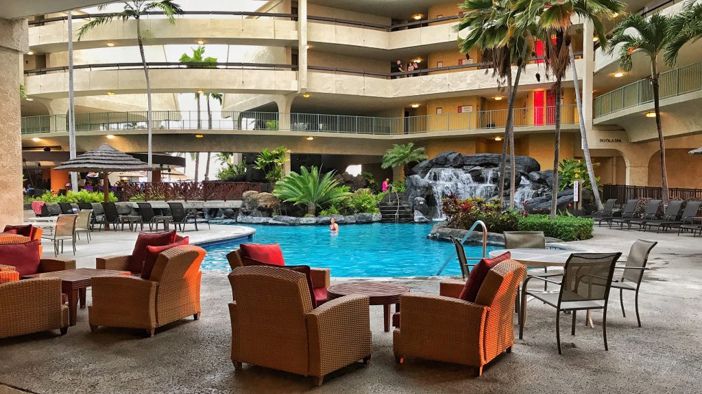 The view of the Sheraton Kona Resort & Spa's poor that is surrounded by palm trees and red-orange couch sofas and a walkways in the background at Keauhou Bay