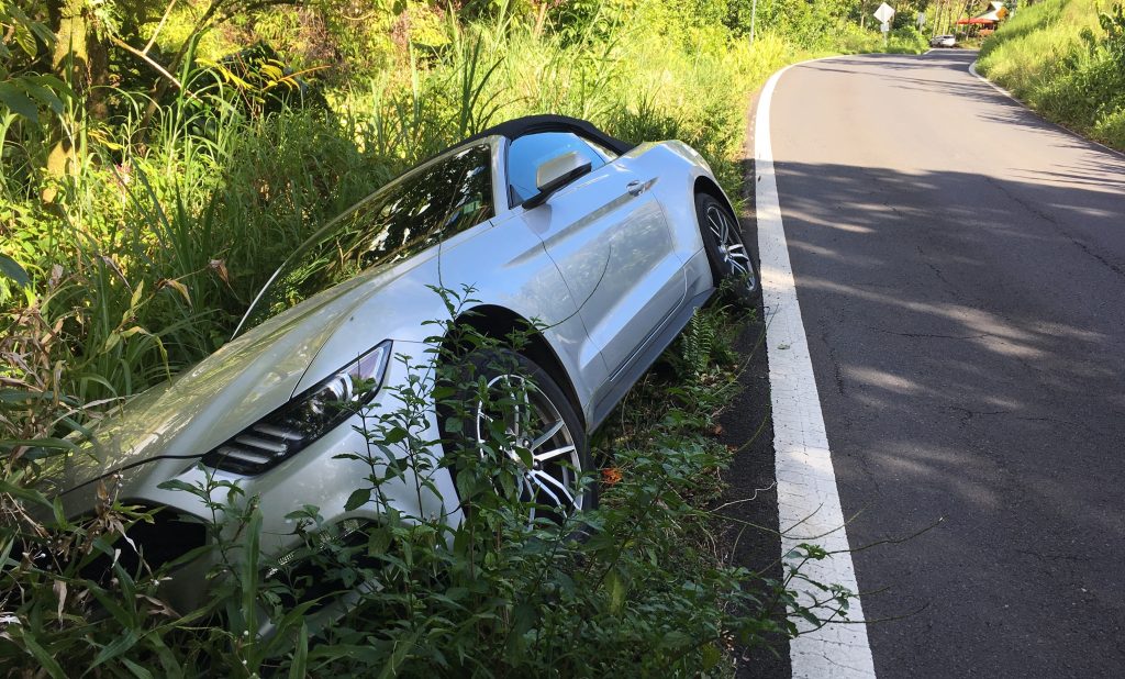 A silver car that's crashed on the side of the road on it's side located in Big Island Hawaii