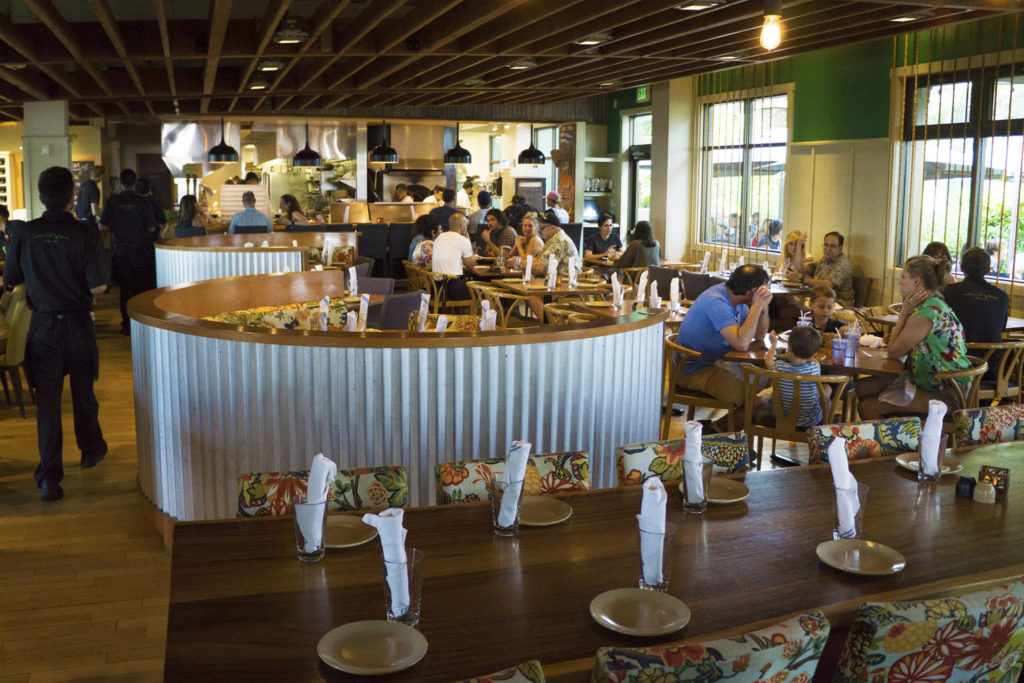 The inside of Monkey Pod Kitchen, in multiple locations in Hawaii, where families are sitting in wooden tables and enjoying their meals.
