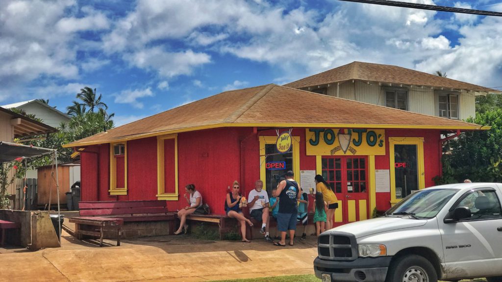 JoJo’s Shave Ice showcasing a red vibrant storefront and surrounded by multiple visitors or customers enjoying refreshing frozen treats amidst the tropical island atmosphere.