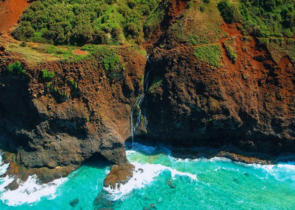 A aerial view of the Kalalau Trail surrounded by nature valleys, a sea cliffs, and a dropdown waterfall into the bright teal body of water in the Na Pali Coast on the island of Kauai, Hawaii.