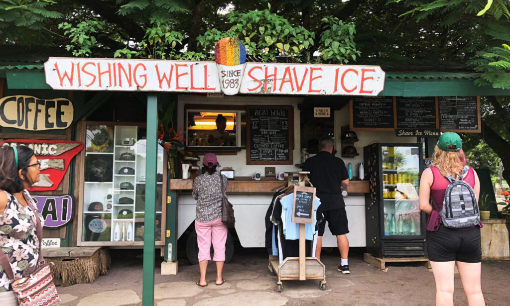 The Wishing Well Shave Ice is a old bus at the entrance to Hanalei town reestablished as a food shack around lush greenery, serving Hawaiian shaved ice treats to multiple customers 