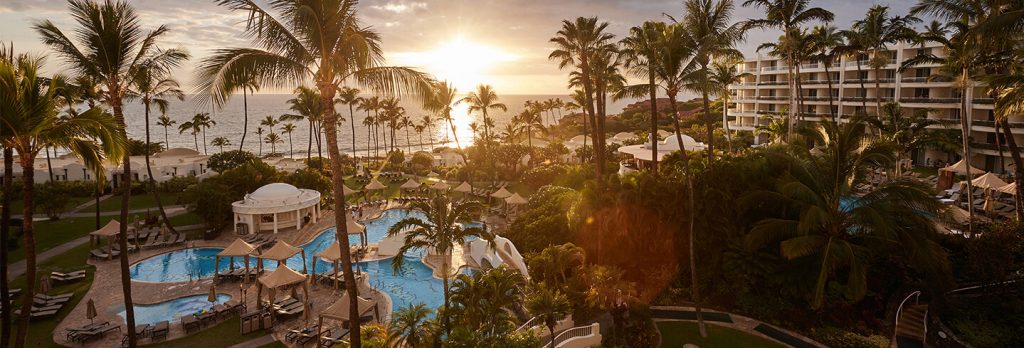 A far shot of Fairmont Kea Lani where you can see a side of it's building, the pool, palm trees and a sun set in the far distance
