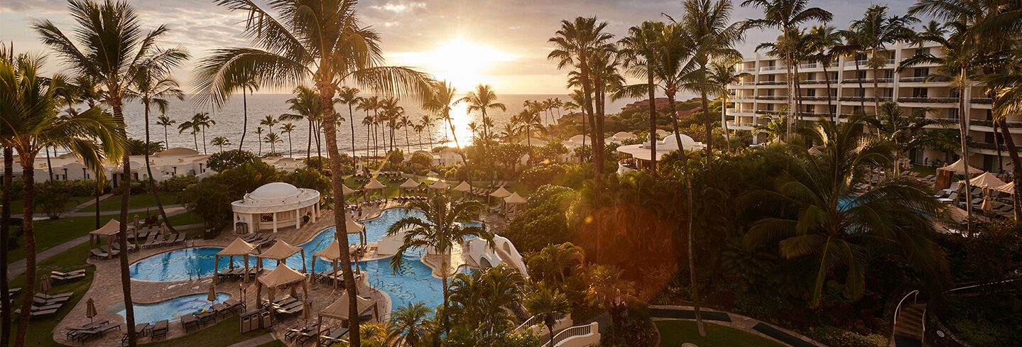 A far shot of Fairmont Kea Lani where you can see a side of it's building, the pool, palm trees and a sun set in the far distance