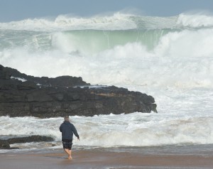 A guy walking on the beach shore while a ocean wave is heading towards the shoreline