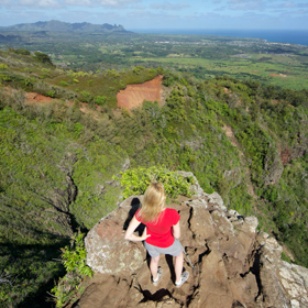 A female hiker with a red shirt is standing at the very end of a trail and looking at the view