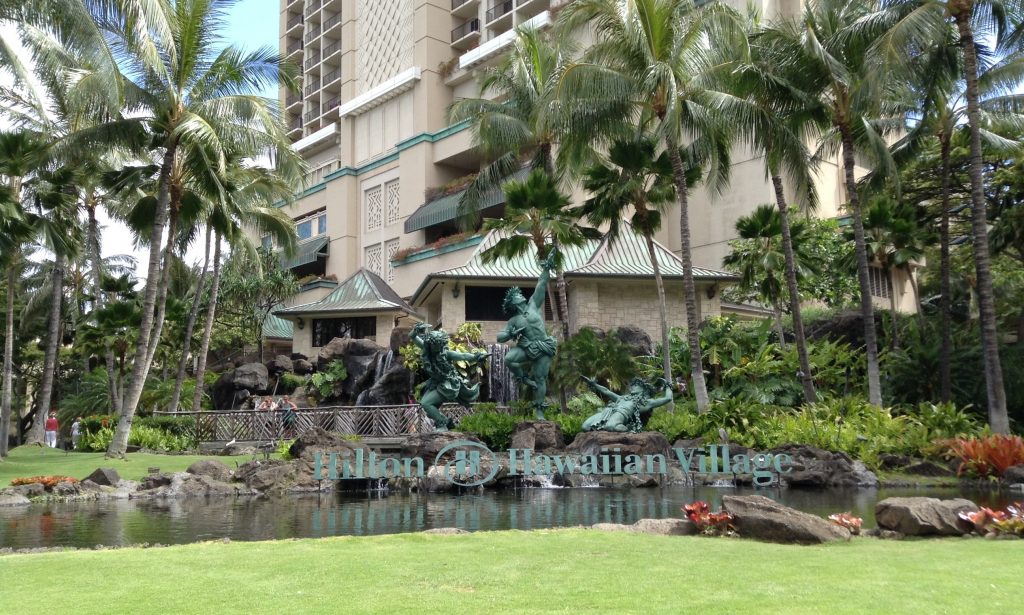 Hilton Hawaiian Village, an oceanfront resort in Honolulu with five towers on Waikiki Beach, with a sign of its name on top of a body of water.